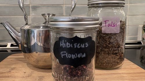 two jars containing herbs labeled Tulsi and Hibiscus flowers. Flanked by a tea kettle